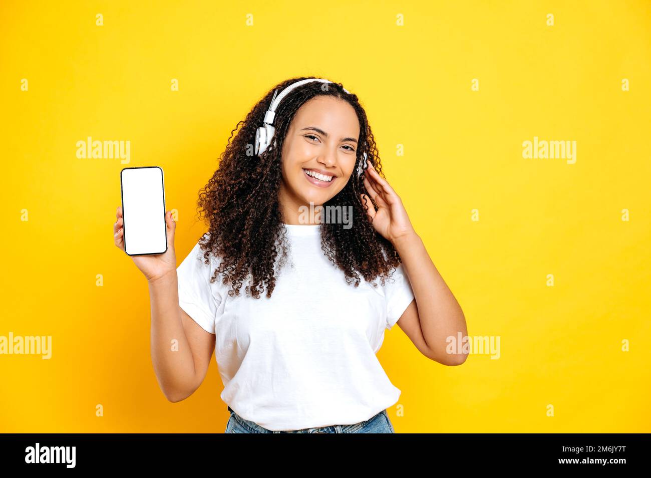 Positive happy lovely brunette brazilian woman, in casual wear, with headphones and cellphone with white mock-up screen, listens her favorite music, dancing, relaxing, smile,isolated yellow background Stock Photo