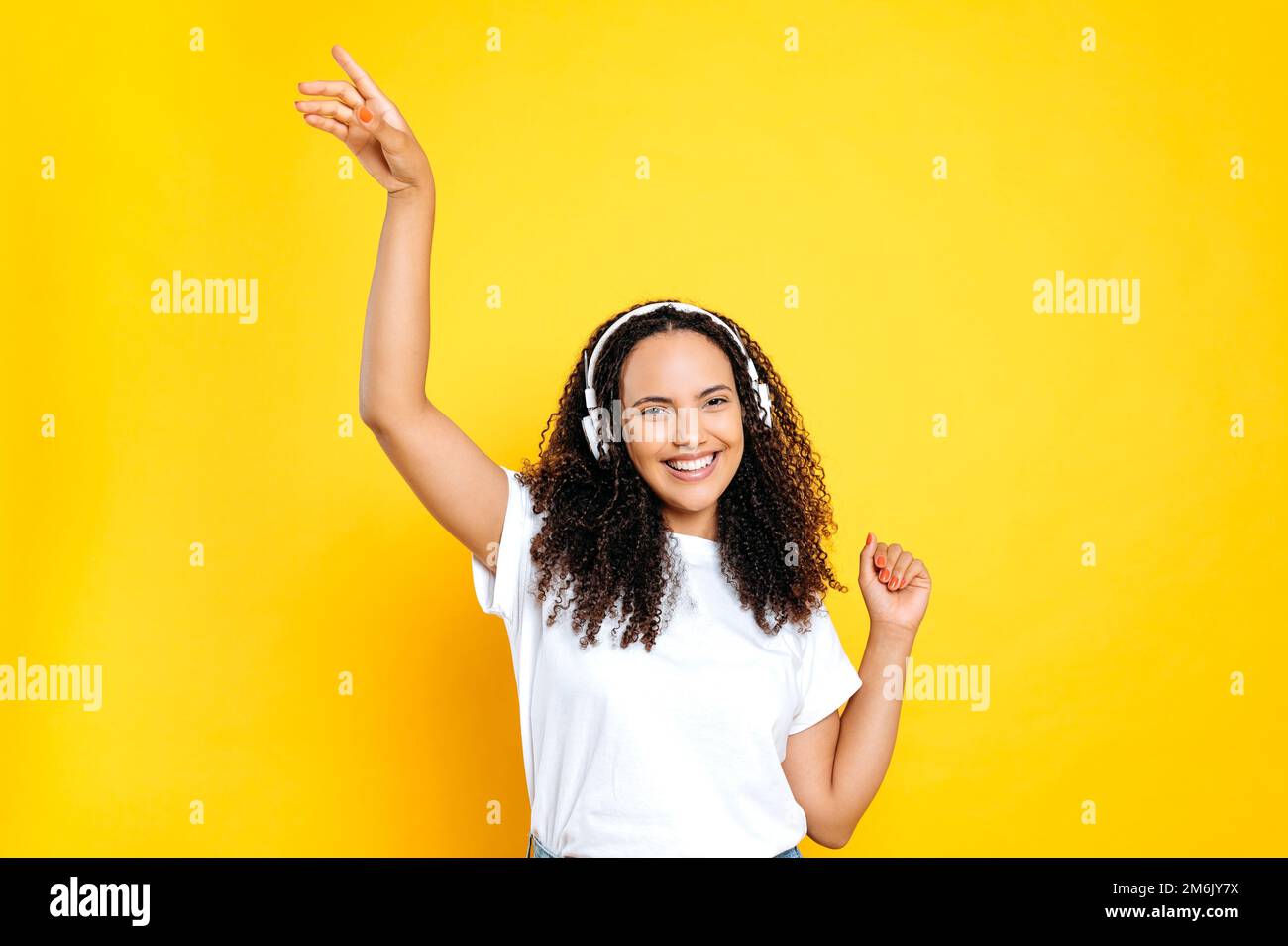 Listen to music, dance. Joyful pretty curly brazilian or latino woman, in casual wear, with headphones, listens her favorite music, dancing relaxing, having fun on isolated yellow background, smiles Stock Photo