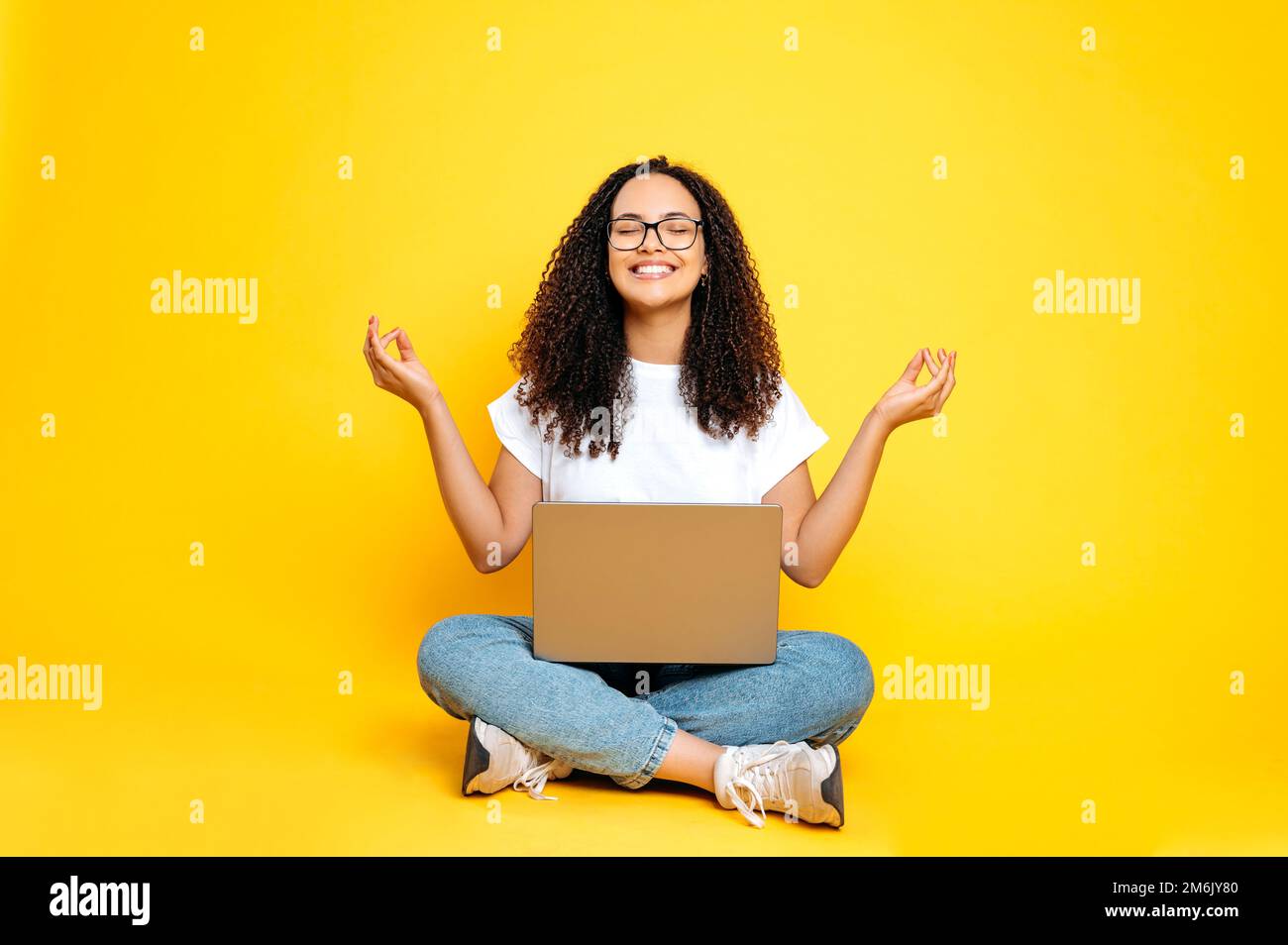 Full-length photo of a positive lovely latino or brazilian woman, in casual wear, sitting with an open laptop in a lotus position with eyes closed on isolated yellow background, relaxing, meditating Stock Photo