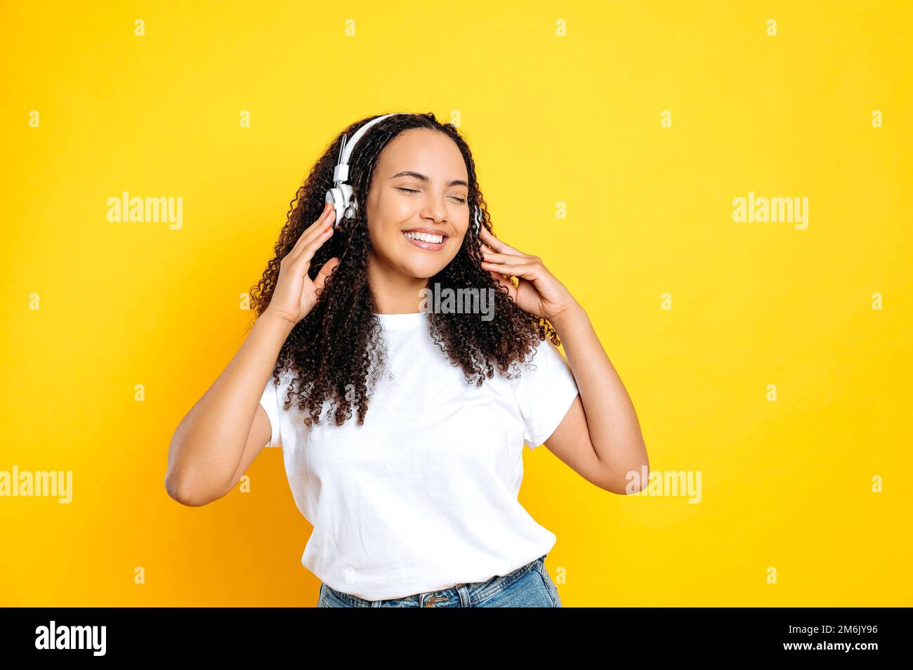 Joyful pretty brunette hispanic woman, in casual basic wear, with headphones, listens her favorite music, dancing with eyes closed, relaxing, having fun on isolated yellow background, smiles Stock Photo