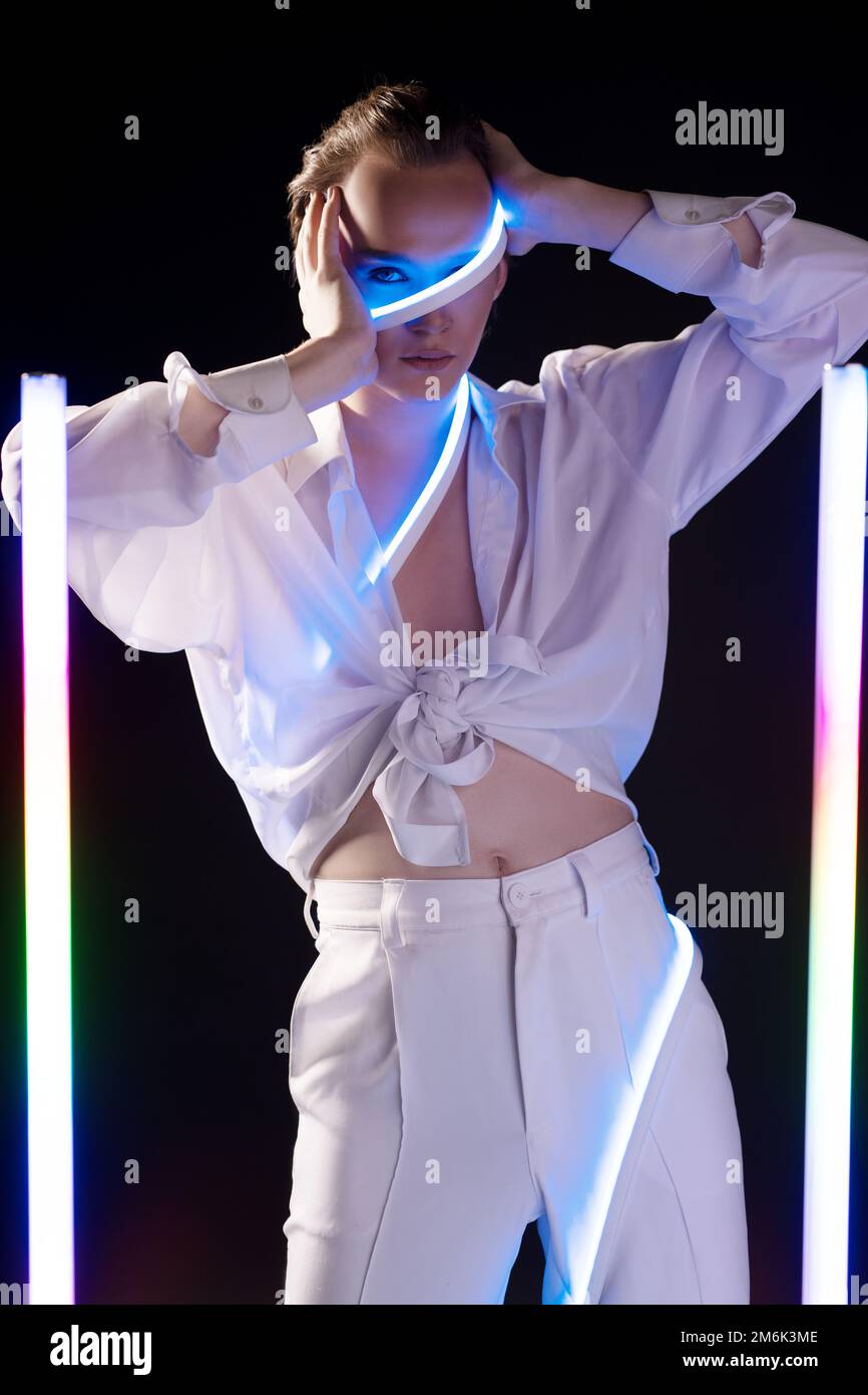 Serious androgynous man standing against black background near neon sticks Stock Photo