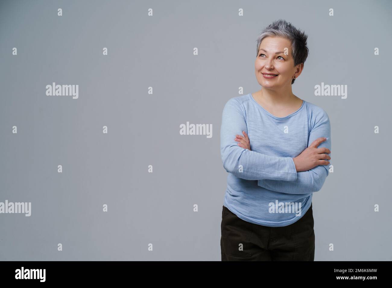 Mature grey hair woman posing with hands folded in blue blouse looking sideways up, copy space on left isolated on white backgro Stock Photo
