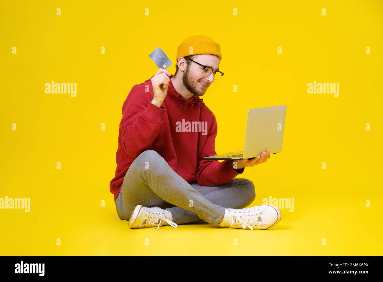 Young man sitting on the floor holding laptop and debit or credit bank card wearing red hoodie and yellow hat buying or paying o Stock Photo