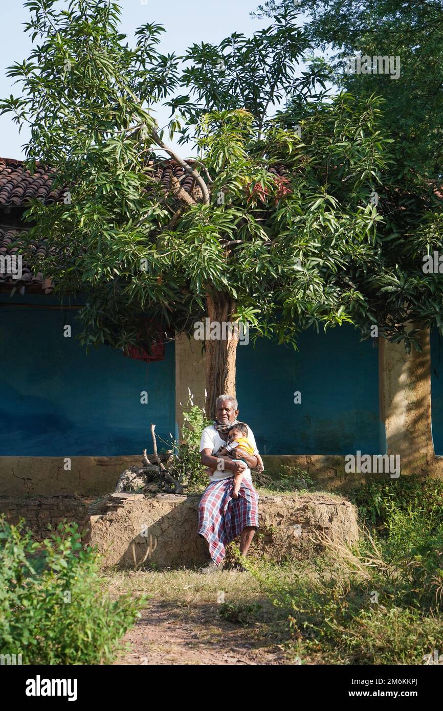 NOVEMBER 15 2022, KACHANDA, CHHATTISGARH, INDIA: Grandfather holding grandson in indian village and sitting under the tree in morning sunlight. Grandf Stock Photo