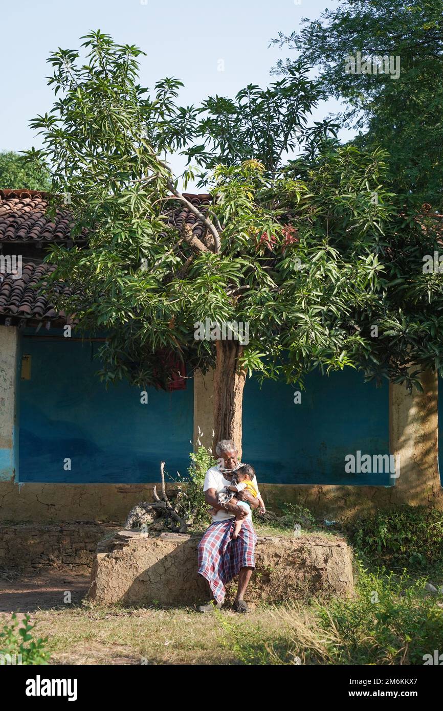 NOVEMBER 15 2022, KACHANDA, CHHATTISGARH, INDIA: Grandfather holding grandson in indian village and sitting under the tree in morning sunlight. Grandf Stock Photo