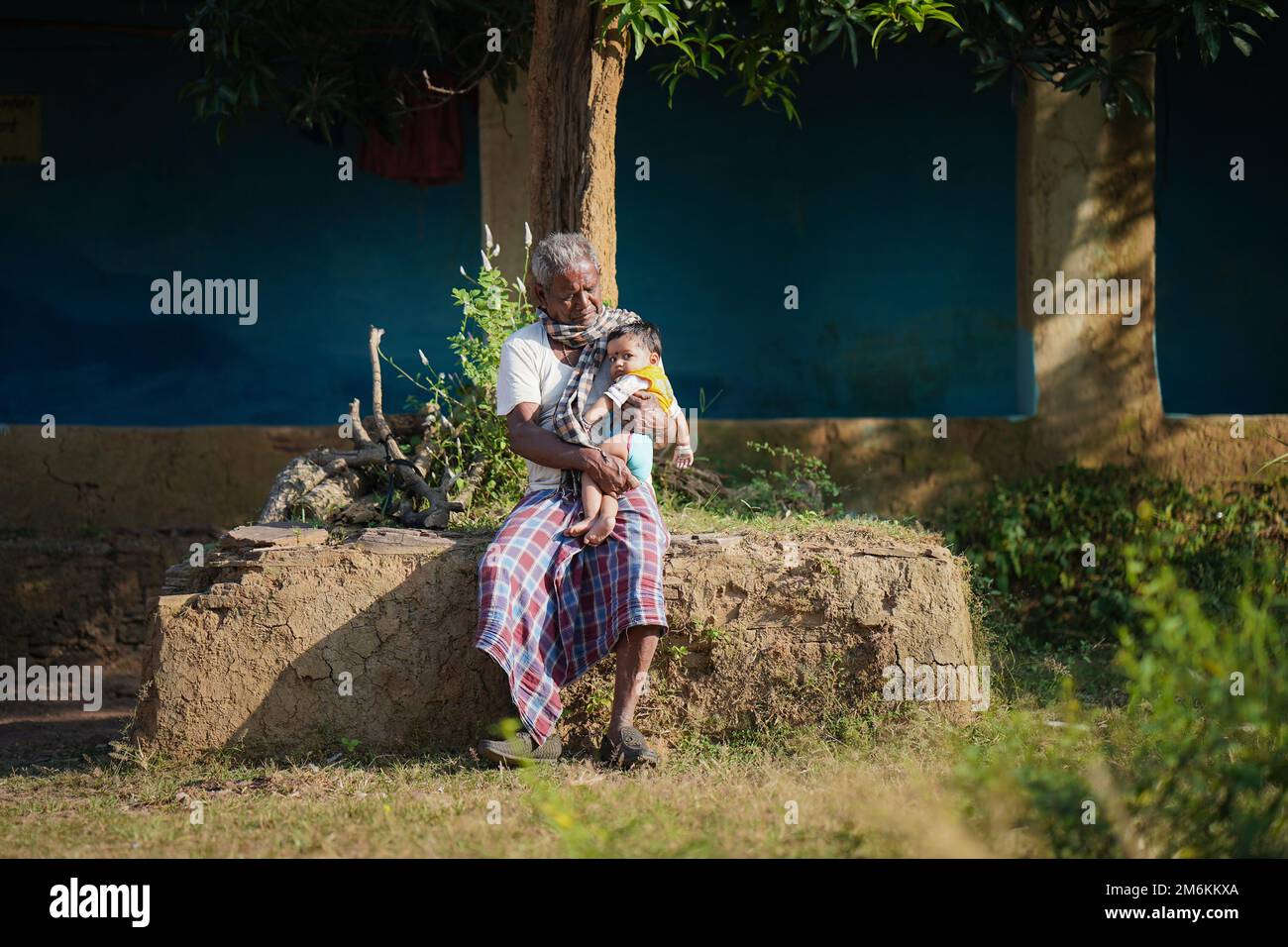 NOVEMBER 15 2022, KACHANDA, CHHATTISGARH, INDIA: Grandfather holding grandson in indian village and sitting under the tree in morning sunlight. Grandf Stock Photo