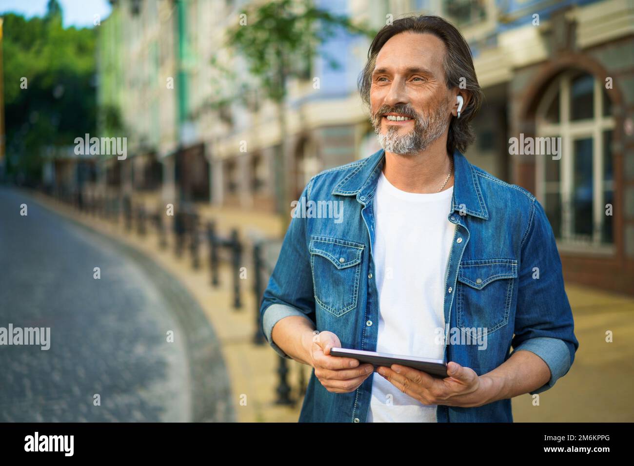 Handsome mature freelancer man with digital tablet in hands having a call while standing on urban streets. Mature man talking or Stock Photo