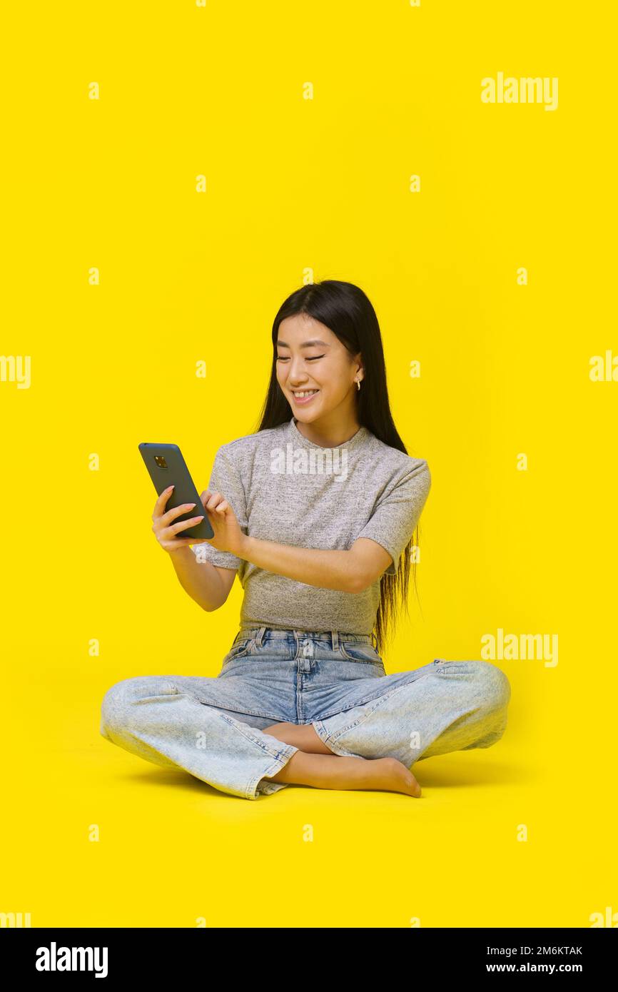 Asian young girl on the floor with phone in hands texting or shopping online playing game isolated on yellow background. Mobile Stock Photo