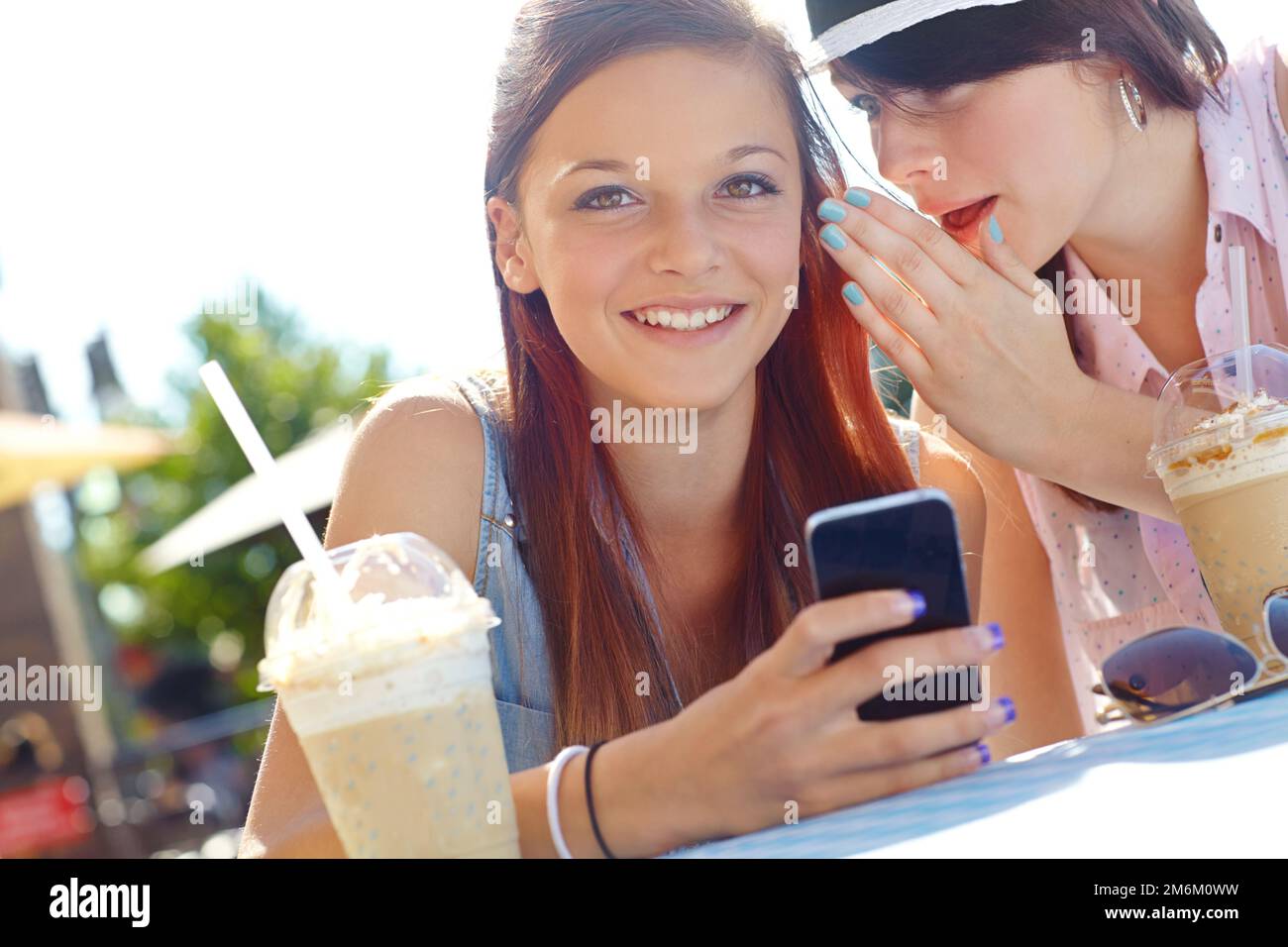 Spread the word...A teenage girl texting on a cellphone while her friend whispers something in her ear. Stock Photo