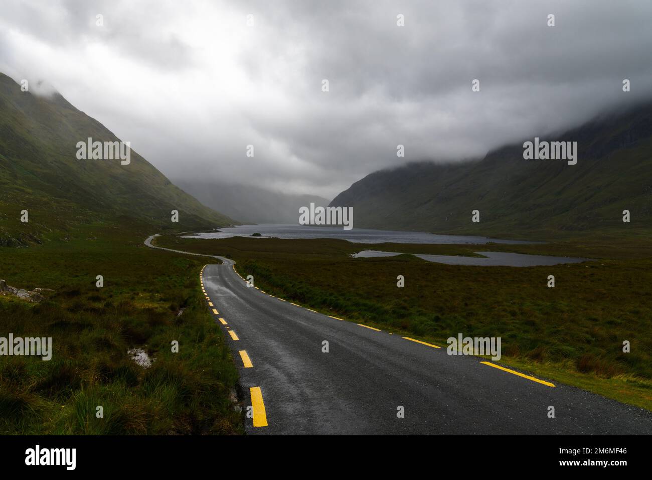 Narrow black-top highway with yellow road markings leads through an overcast mountain valley with fog and mist and lakes o nthe Stock Photo