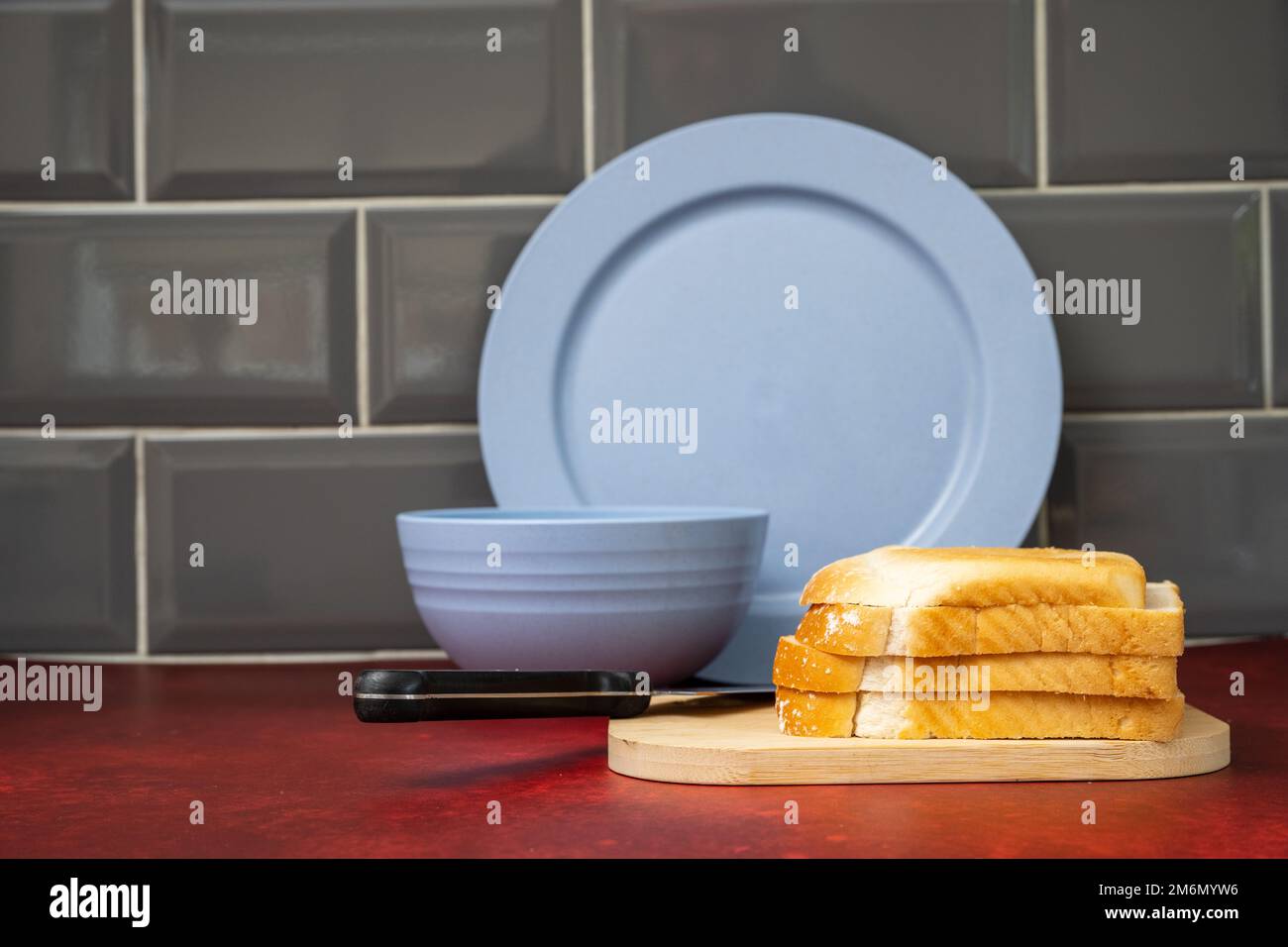 blue pottery china  dinner plate bowl  black handled stainless steel knife on wooden board with fresh bread on red linoleum work top with grey tiled s Stock Photo