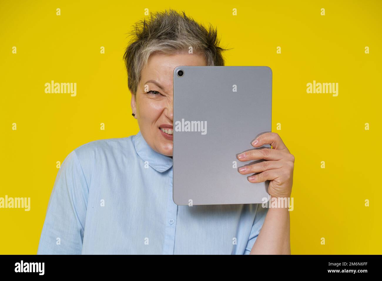 Online troll mature woman hide behind digital tablet with evil facial expression isolated on yellow background. Modern senior wo Stock Photo