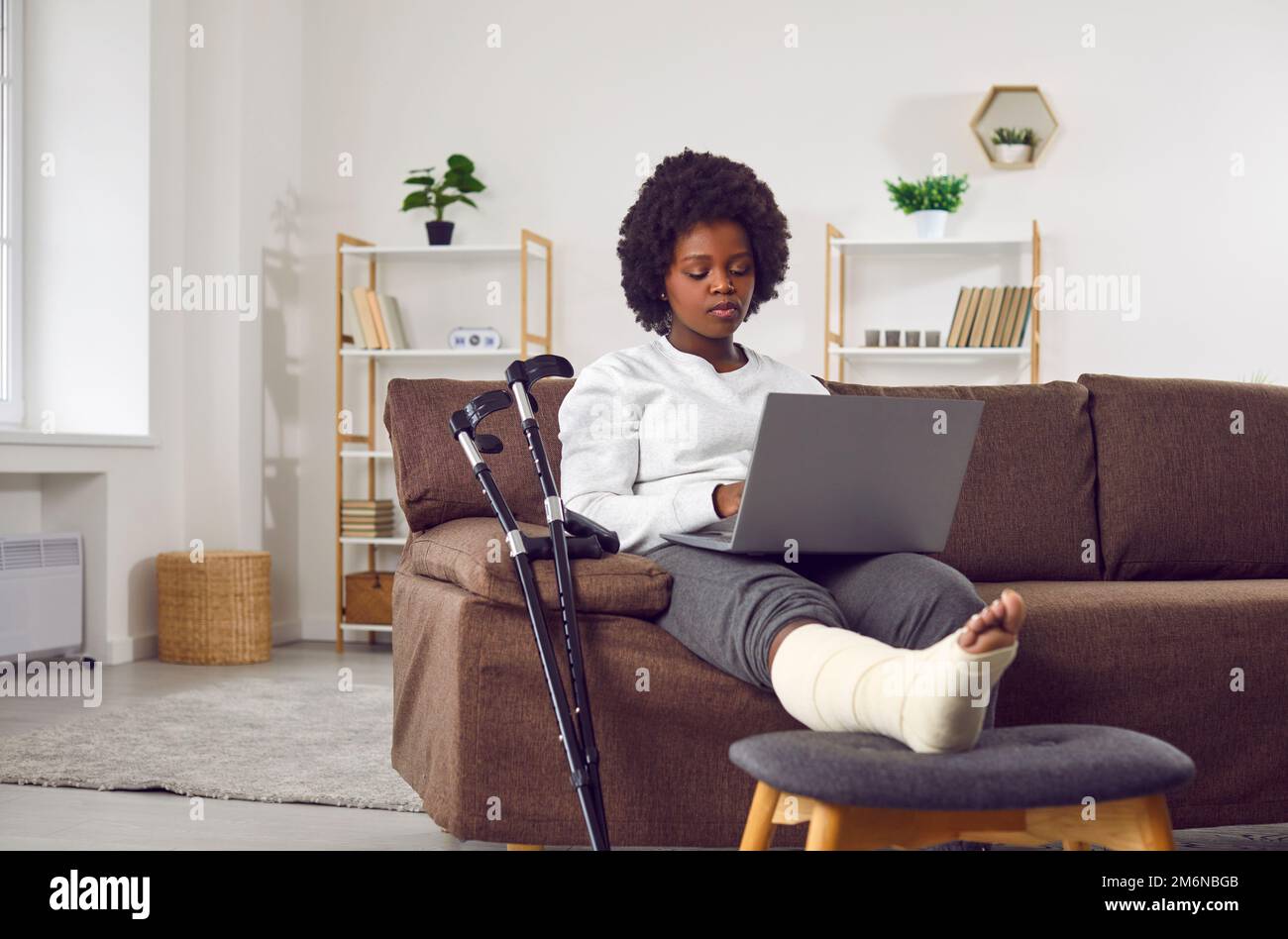 Attractive African American woman with broken leg in cast is sitting on the sofa in the living room with laptop in hands. Stock Photo