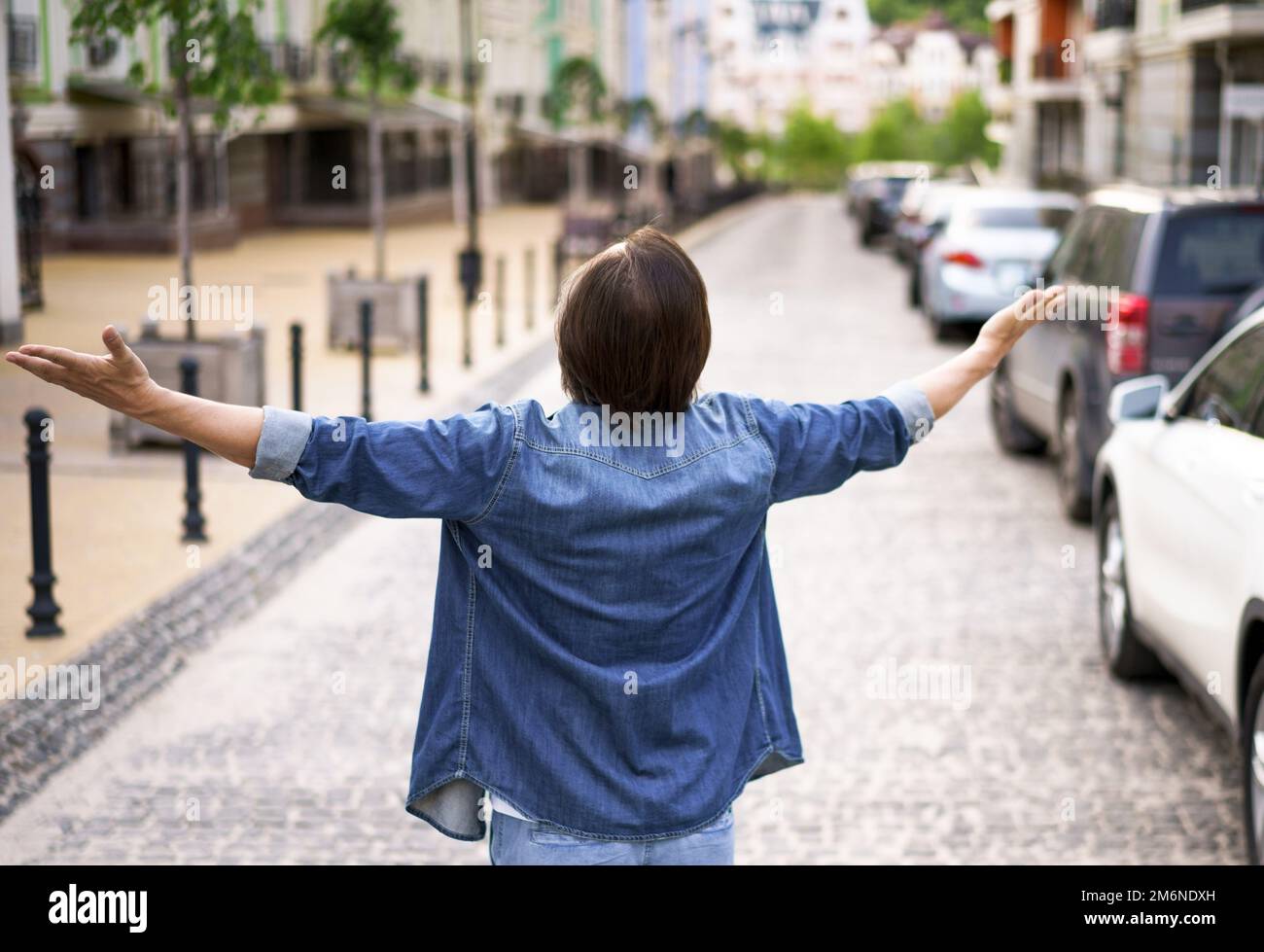 Back view of senior man standing outdoors on the street with hands spread wide open wearing denim jeans shirt. Mid Aged man glad Stock Photo