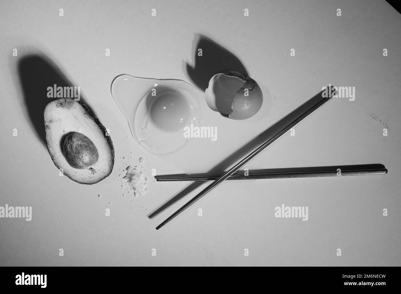 A black and white top view of an avocado, a cracked egg and sticks