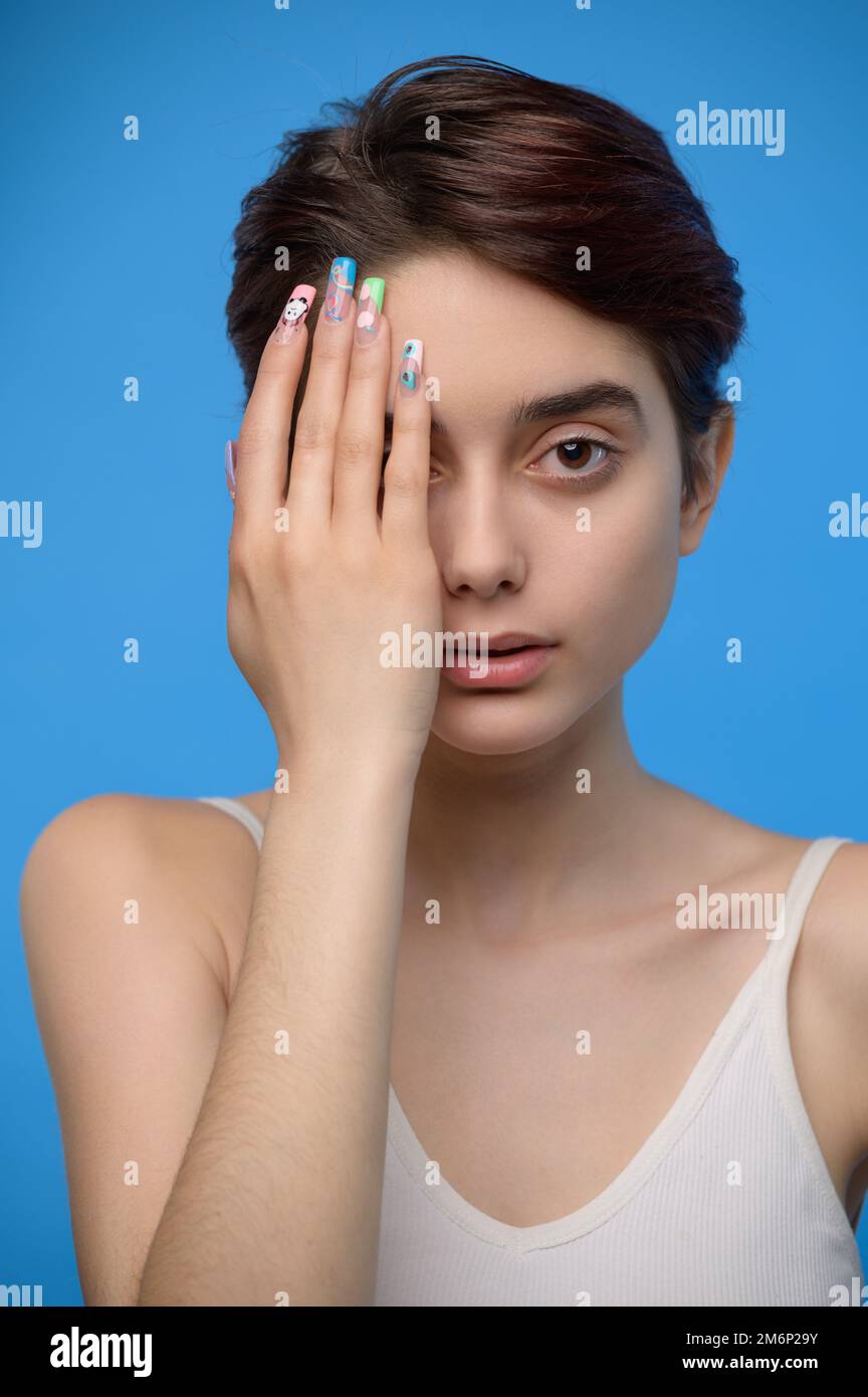 Skinny teenage girl cute girl hiding right half of her face with hand, at blue background Stock Photo