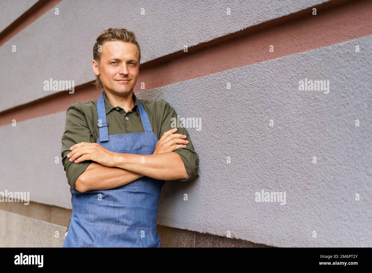 Handsome employee man standing outside with arms folded wearing apron take a break leaned on a wall enjoying fresh air after lon Stock Photo