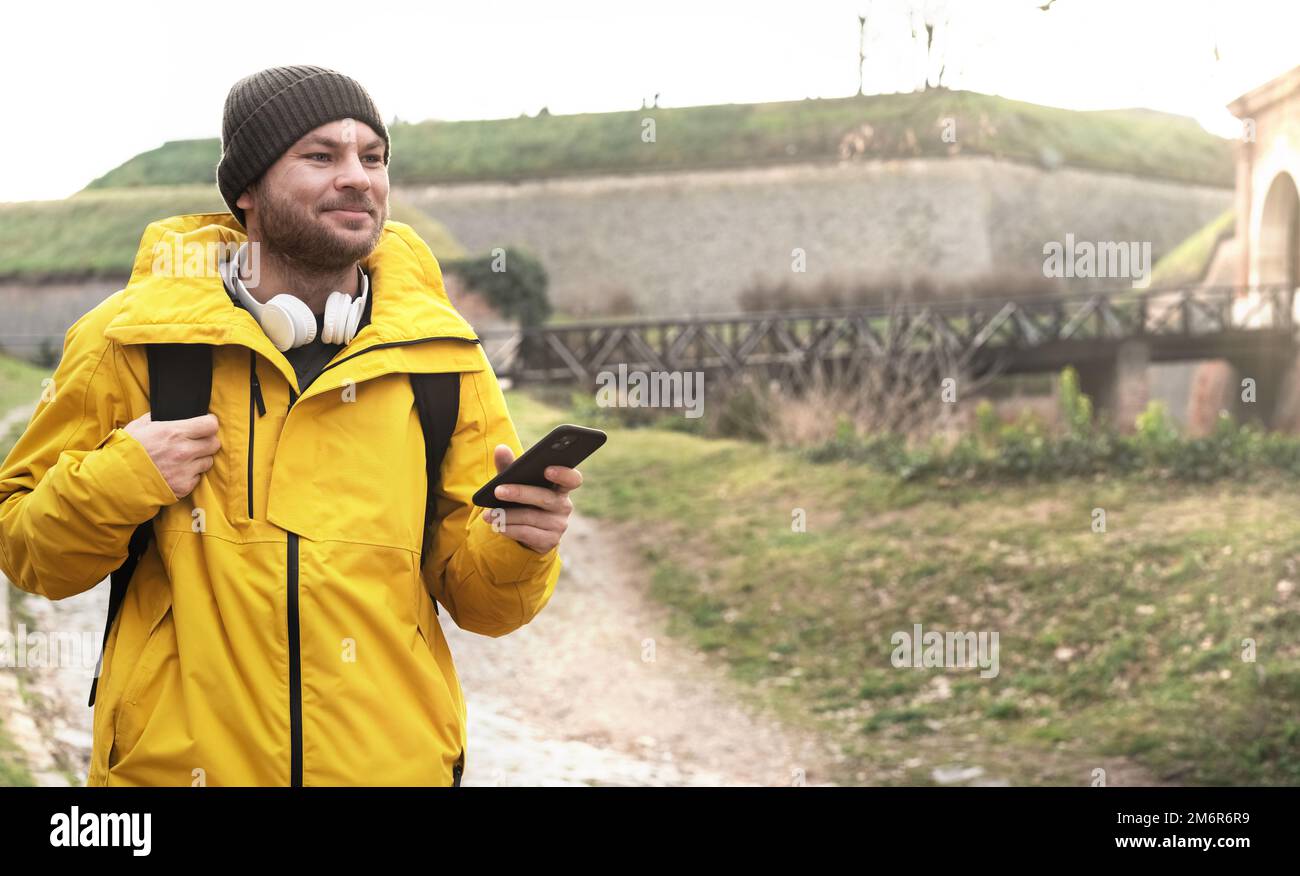 Man in yellow travel jacket with headphones on his neck and smart phone in hand walking around old town. Stock Photo