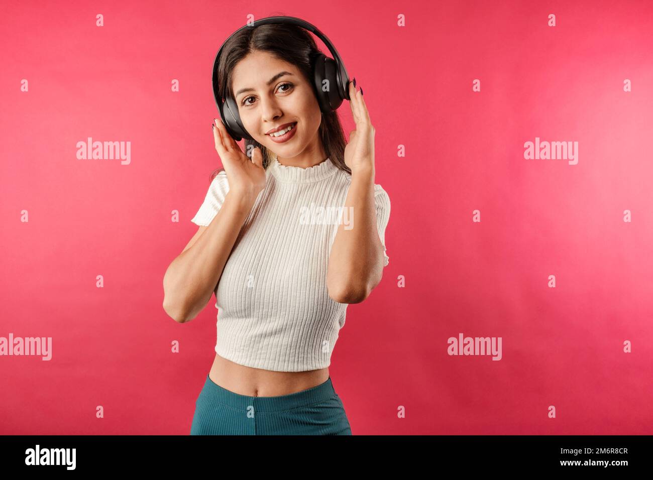 Young beautiful woman wearing ribbed crop isolated over red background listening to music in a pleasant way poses by touching headphones. Very happy, Stock Photo
