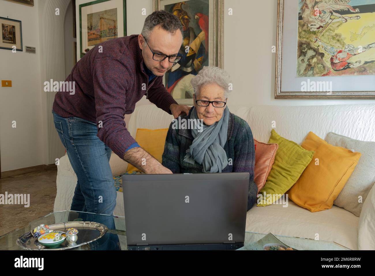 Man helping an old woman with the laptop. Woman sitting on the couch in her living room with a laptop on the table in front of her. Stock Photo