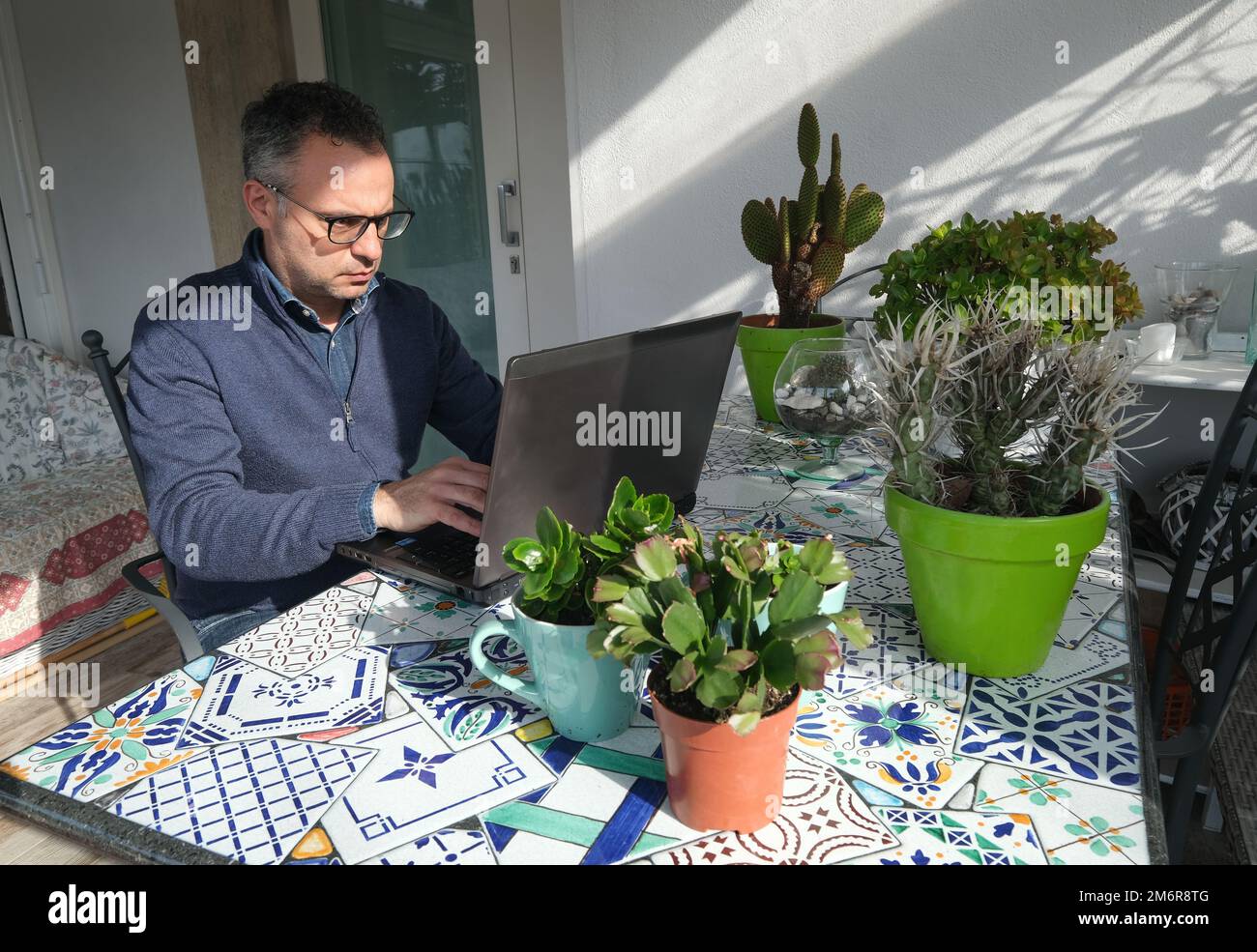 Male working from home in stylish casual clothing with laptop at outdoors veranda with green plants on the table. Italy Stock Photo