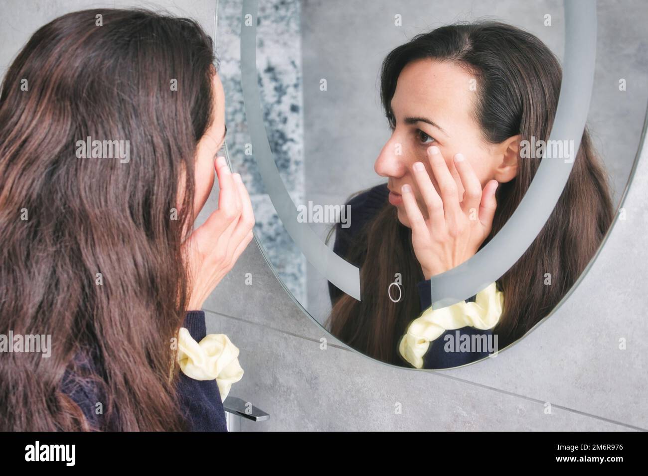 An attractive white Caucasian young woman looking in the bathroom mirror touching her wrinkles around her eyes Stock Photo