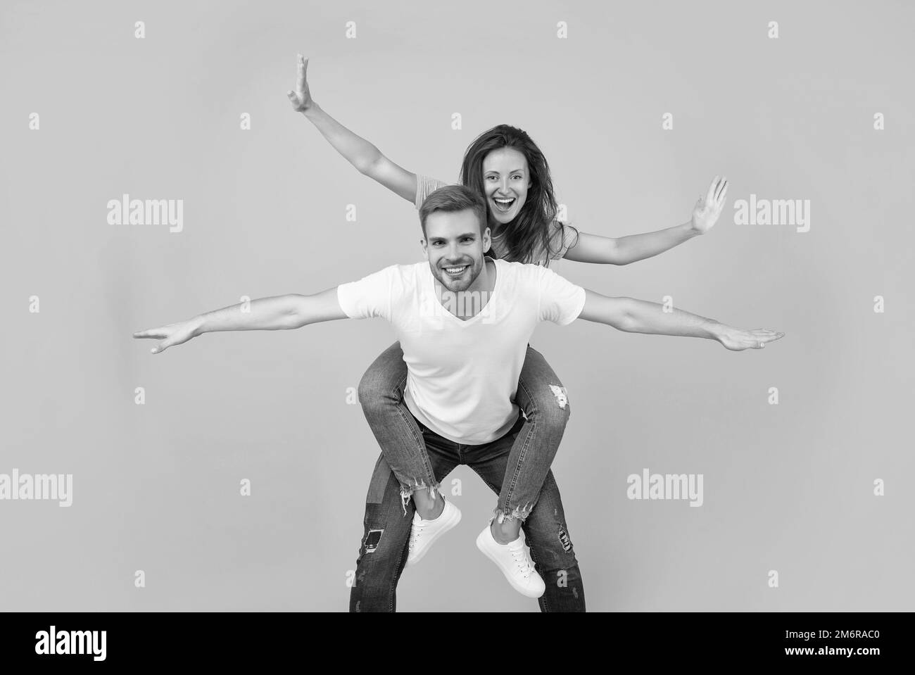 Happy couple in love of girl and man pretend flying doing piggyback ride yellow background, playful Stock Photo