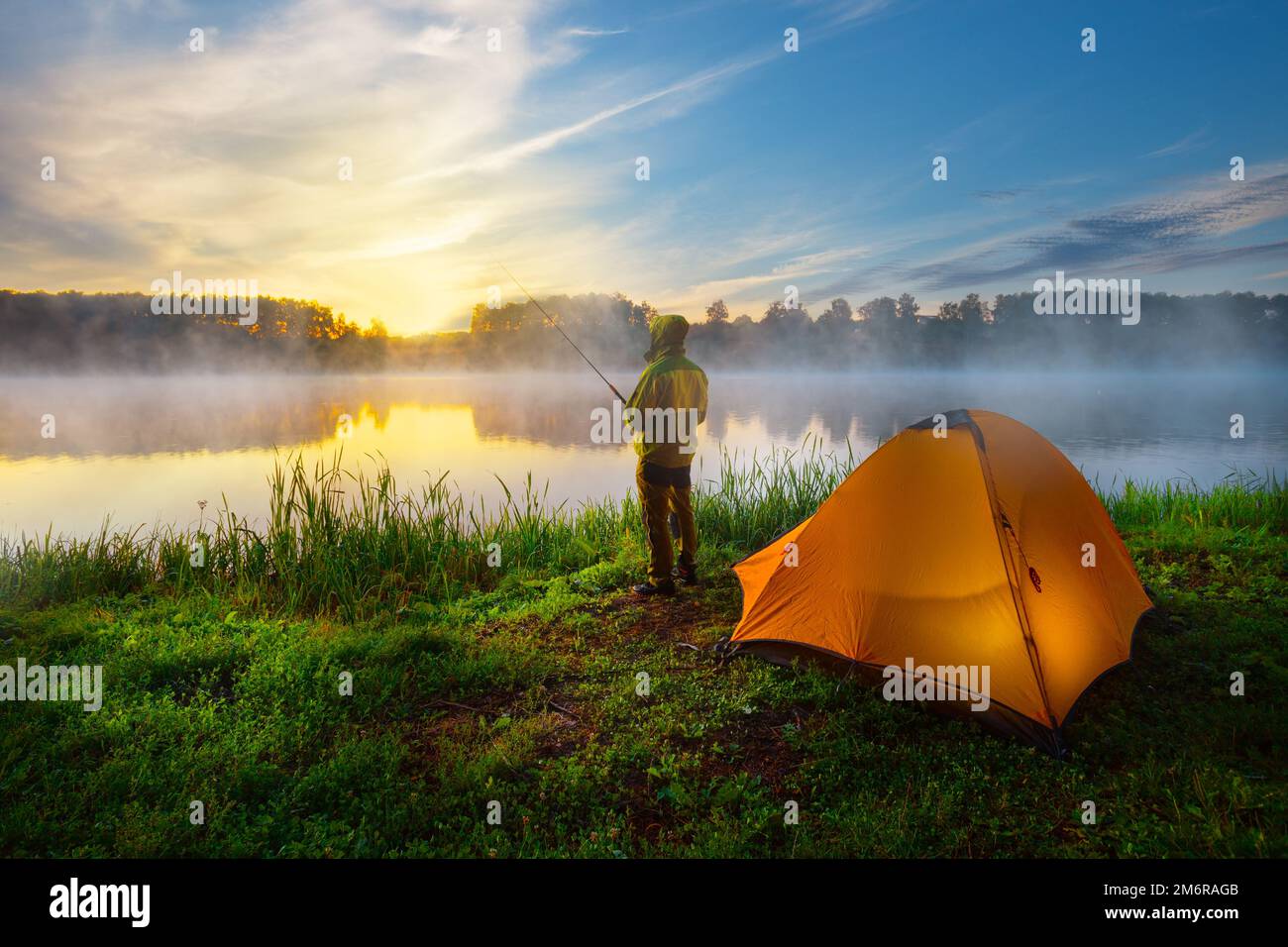 Fisherman on bank of foggy river near an orange tent in the early morning Stock Photo