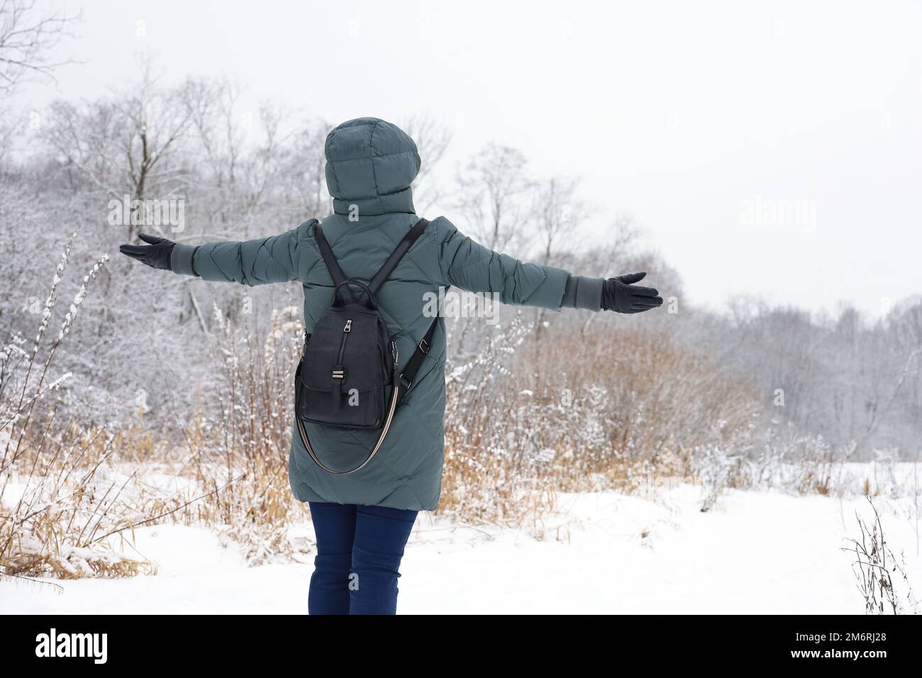 Woman wearing down jacket standing with her hands spread out to the sides and enjoying the snowy weather. Leisure in winter park, cold season Stock Photo