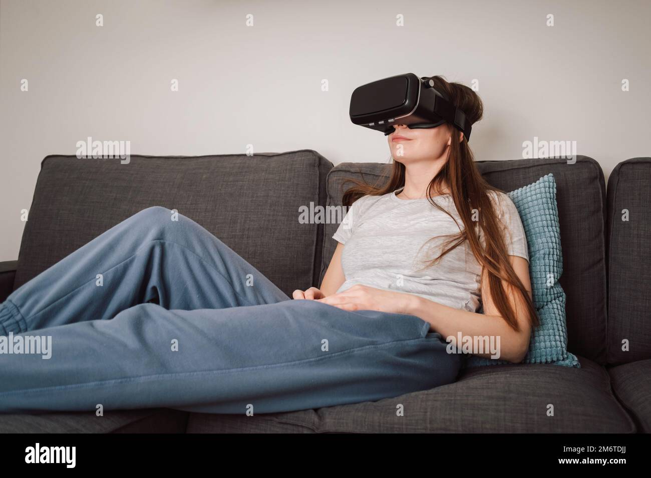 Beautiful smiling young woman wearing VR goggles and virtually traveling through the metaverse from the comfort of her couch Stock Photo