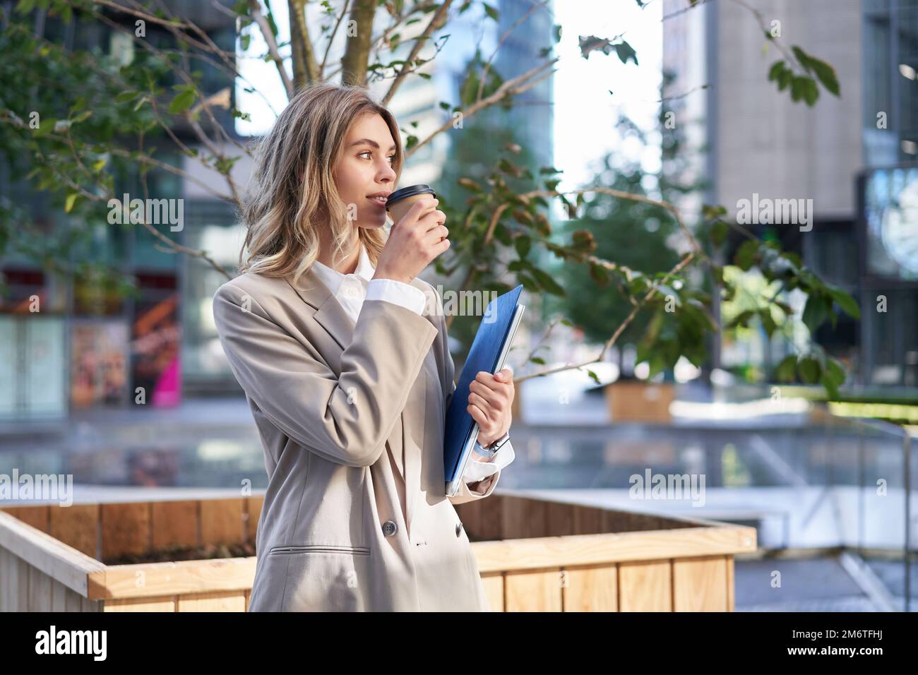 Beautiful smiling businesswoman drinks coffee on her working break, standing on street Stock Photo