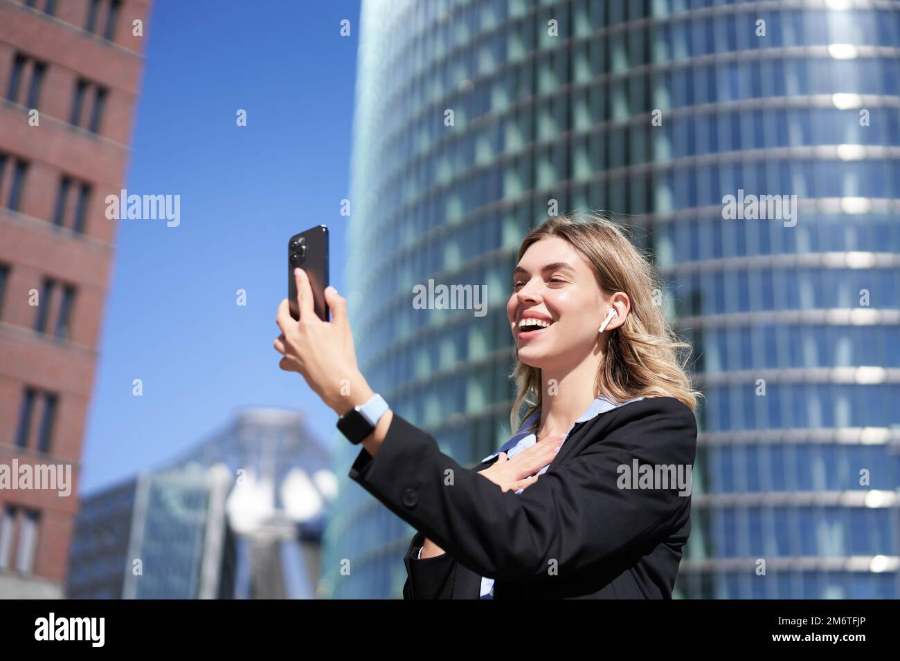 Portrait of smiling corporate woman video call on street, holding mobile phone and waving at smartphone camera, wearing suit Stock Photo