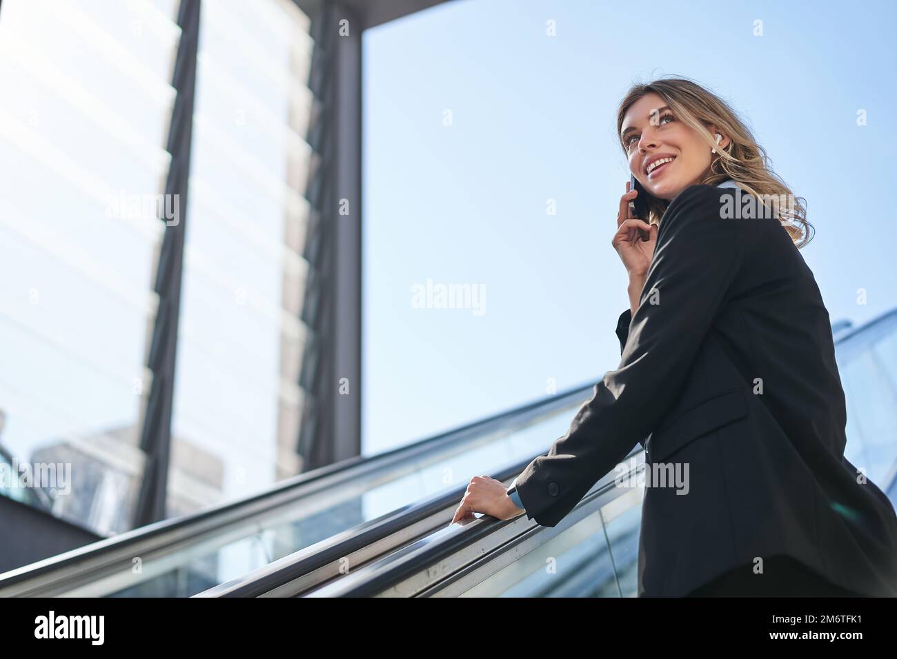 Portrait of businesswoman in black suit, going up on escalator, talking on mobile phone. Saleswoman walking in city, having a ca Stock Photo