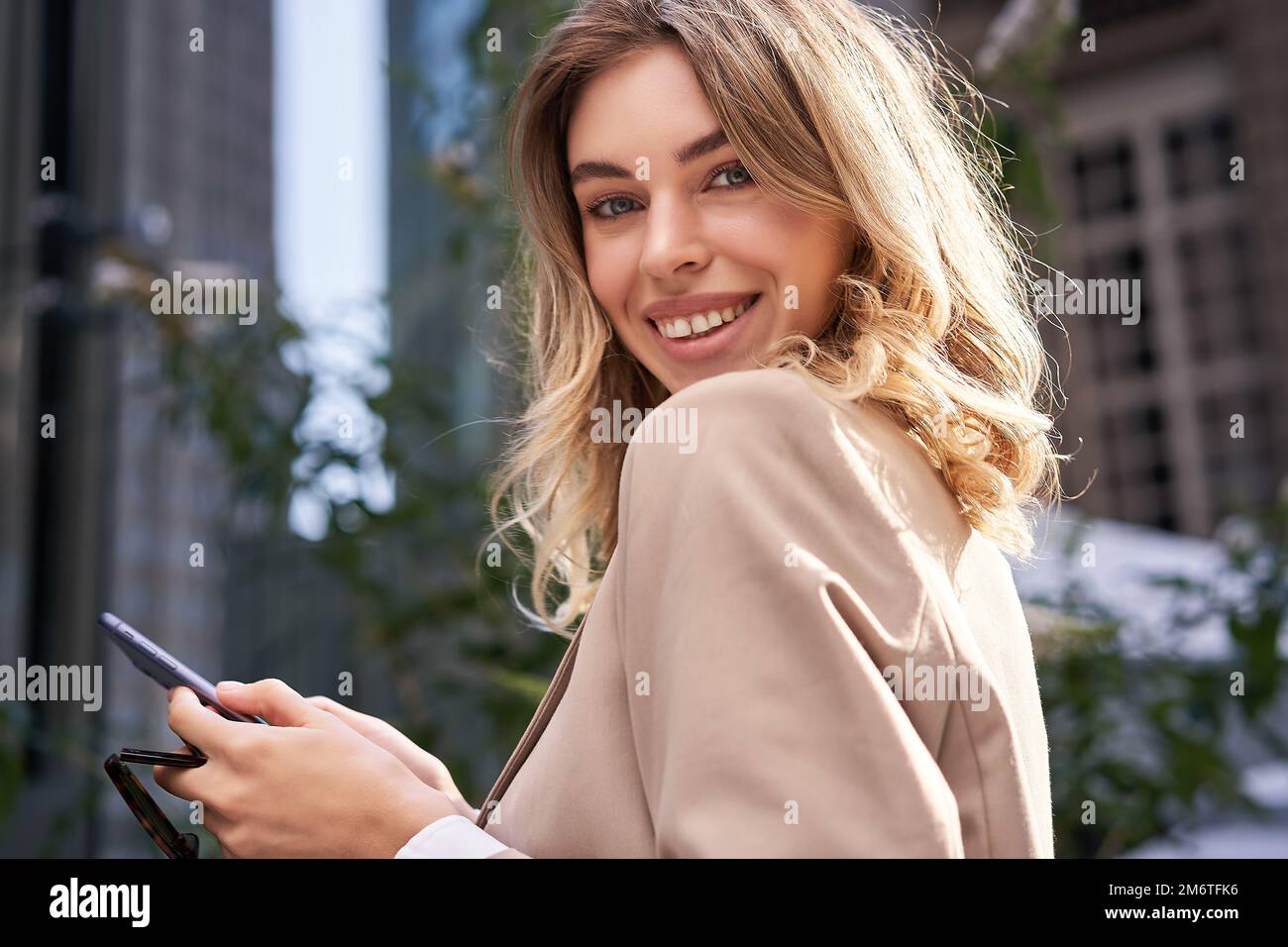 Close up portrait of successful young woman, corporate lady in suit, holds her mobile phone, turns at camera with happy smile, s Stock Photo