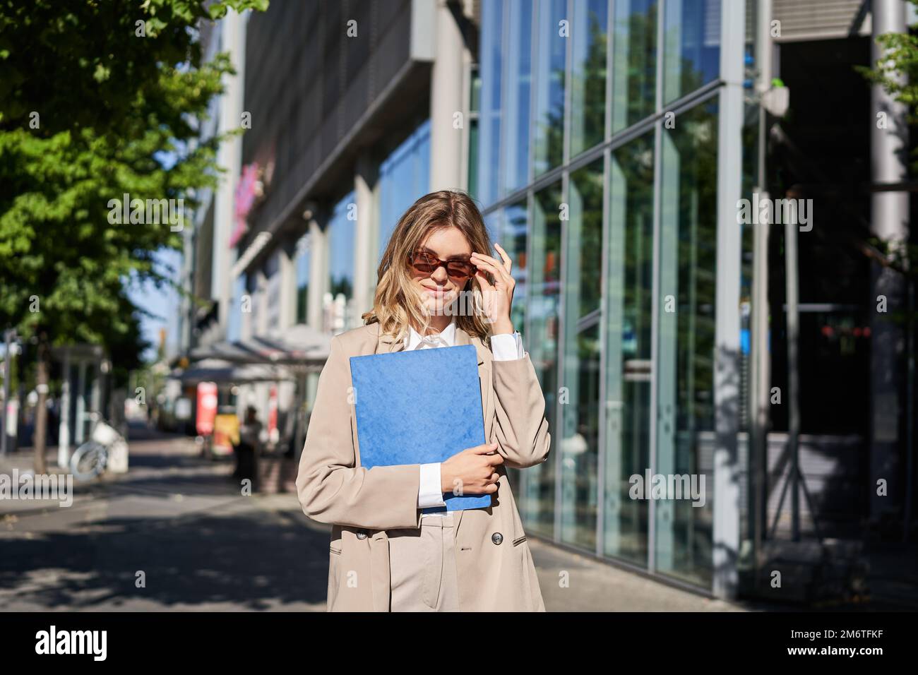 Portrait of corporate woman in sunglasses and beige suit, holding blue folder with office documents, going to work, walking on s Stock Photo