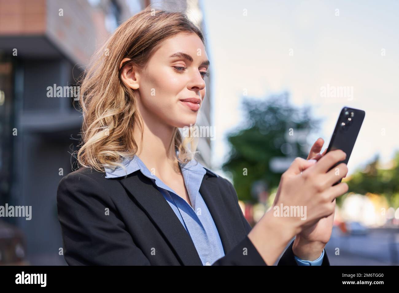 Corporate woman stands on street and texts message on mobile phone, smiling while looking at smartphone screen Stock Photo