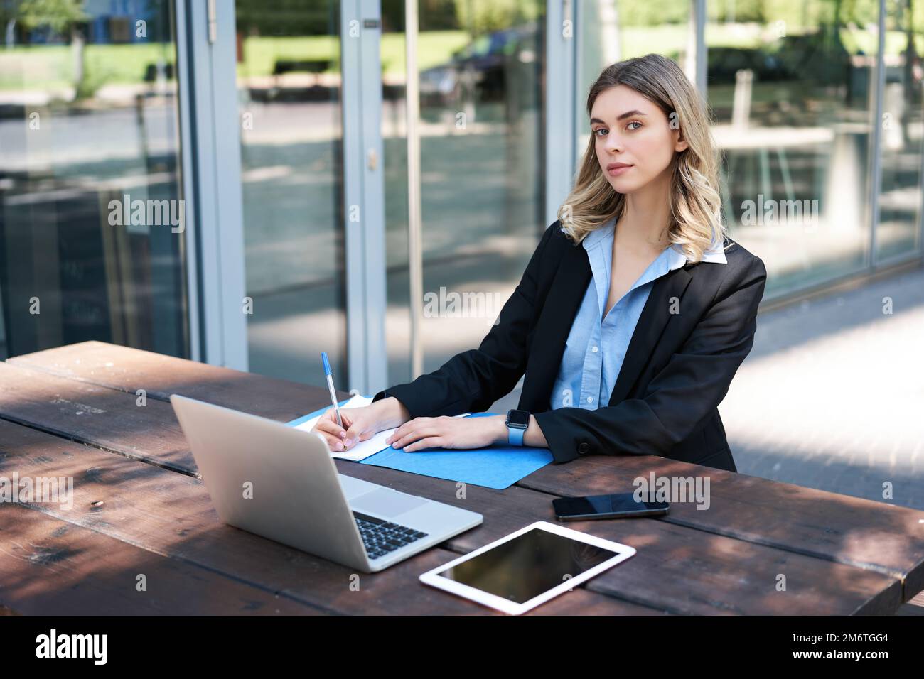 Successful businesswoman working outdoors. Corporate woman sitting on bench with laptop, writing, taking notes during work meeti Stock Photo