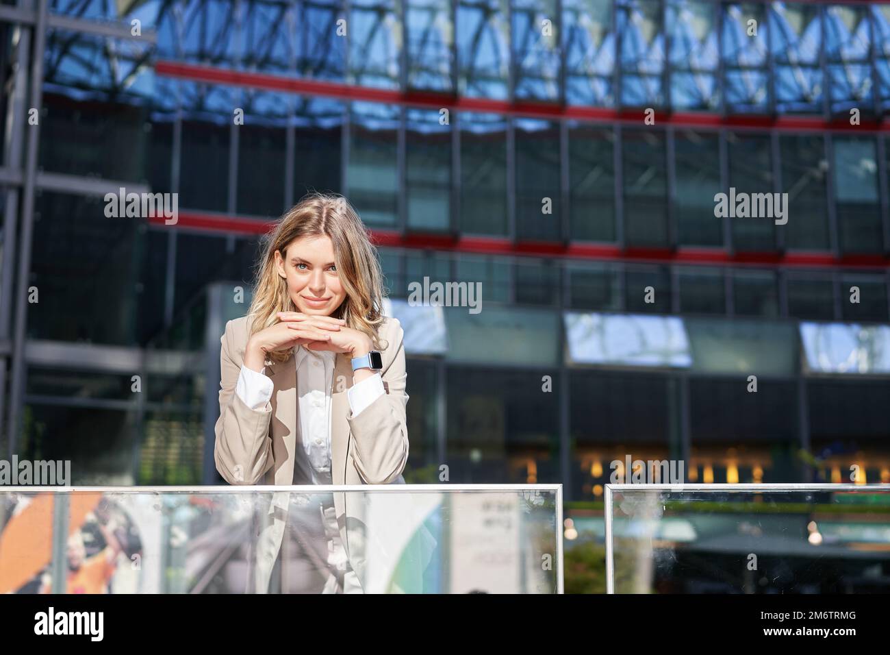 Beautiful young woman in beige suit, standing near office buildings in city center, smiling and looking dreamy Stock Photo