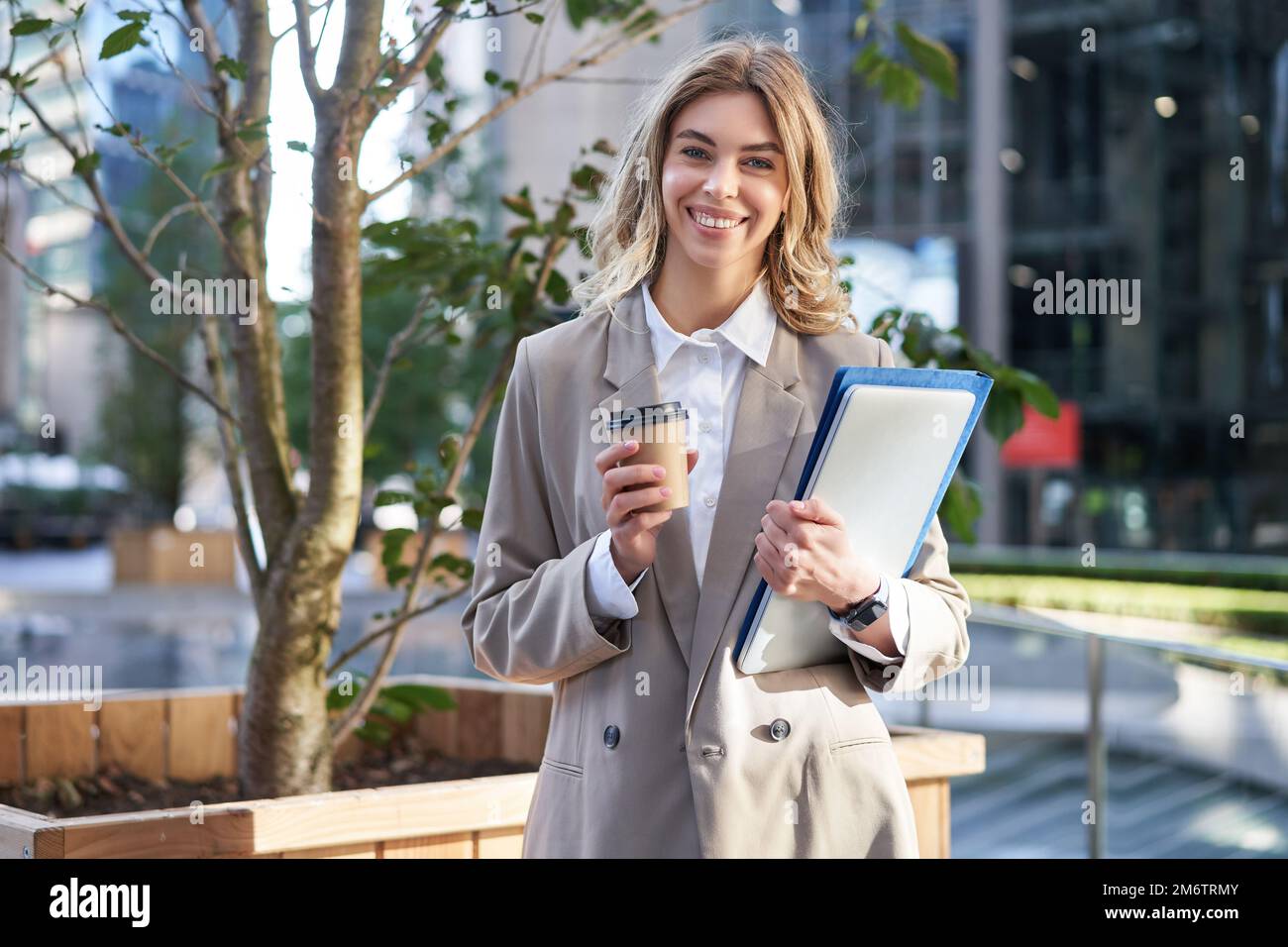 Young businesswoman standing on street with takeaway coffee, laptop and work documents, smiling Stock Photo