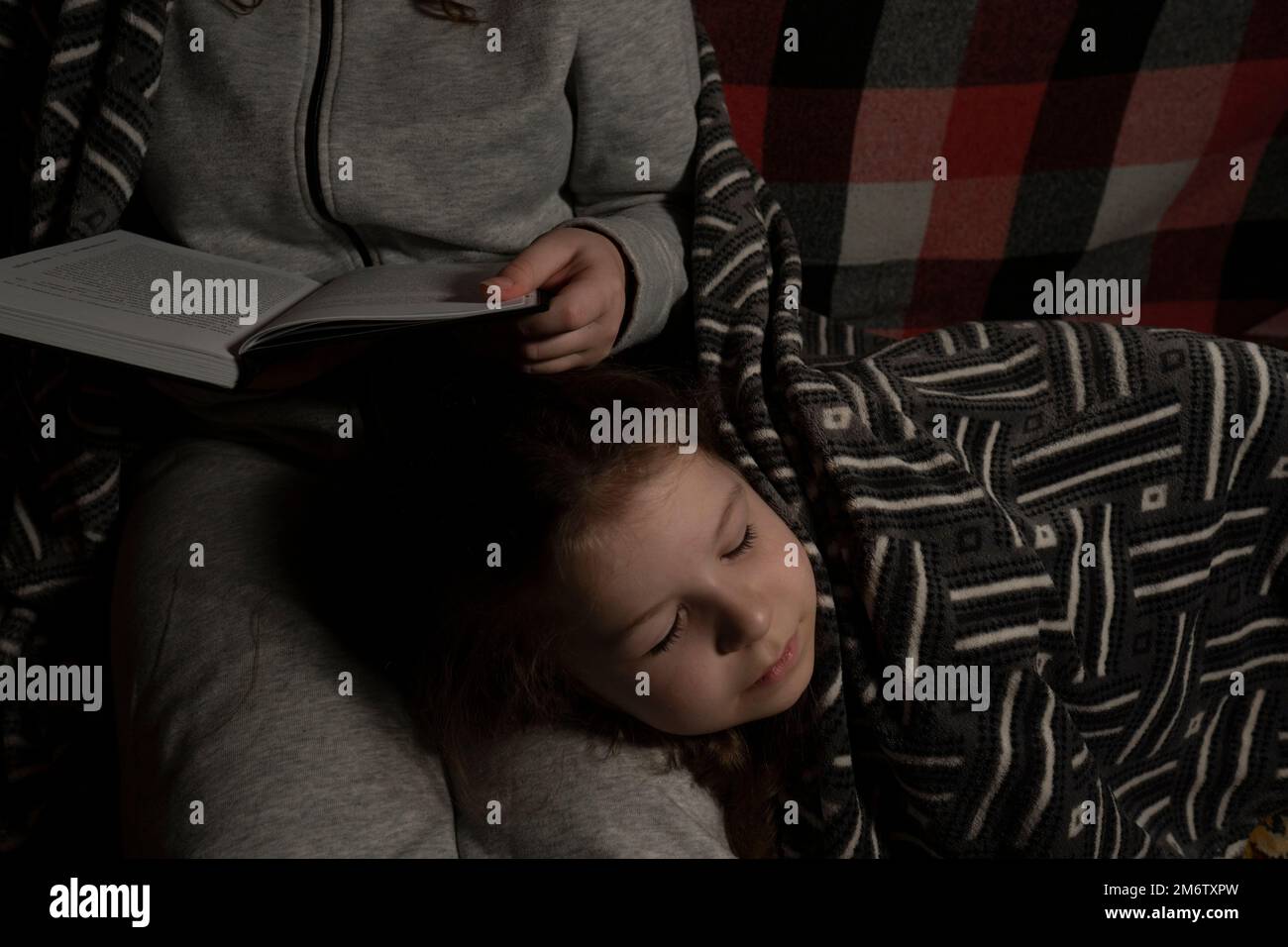 A woman in a dark room reads a book to a sleeping little girl. Blackout. Energy crisis. Destruction of infrastructure. Power outage concept Stock Photo