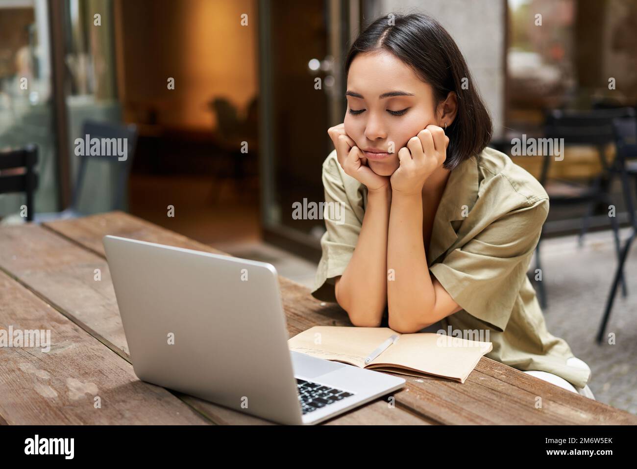Bored girl looks at her laptop, sits outdoors in cafe, listening boring online meeting, working and feeling sad Stock Photo