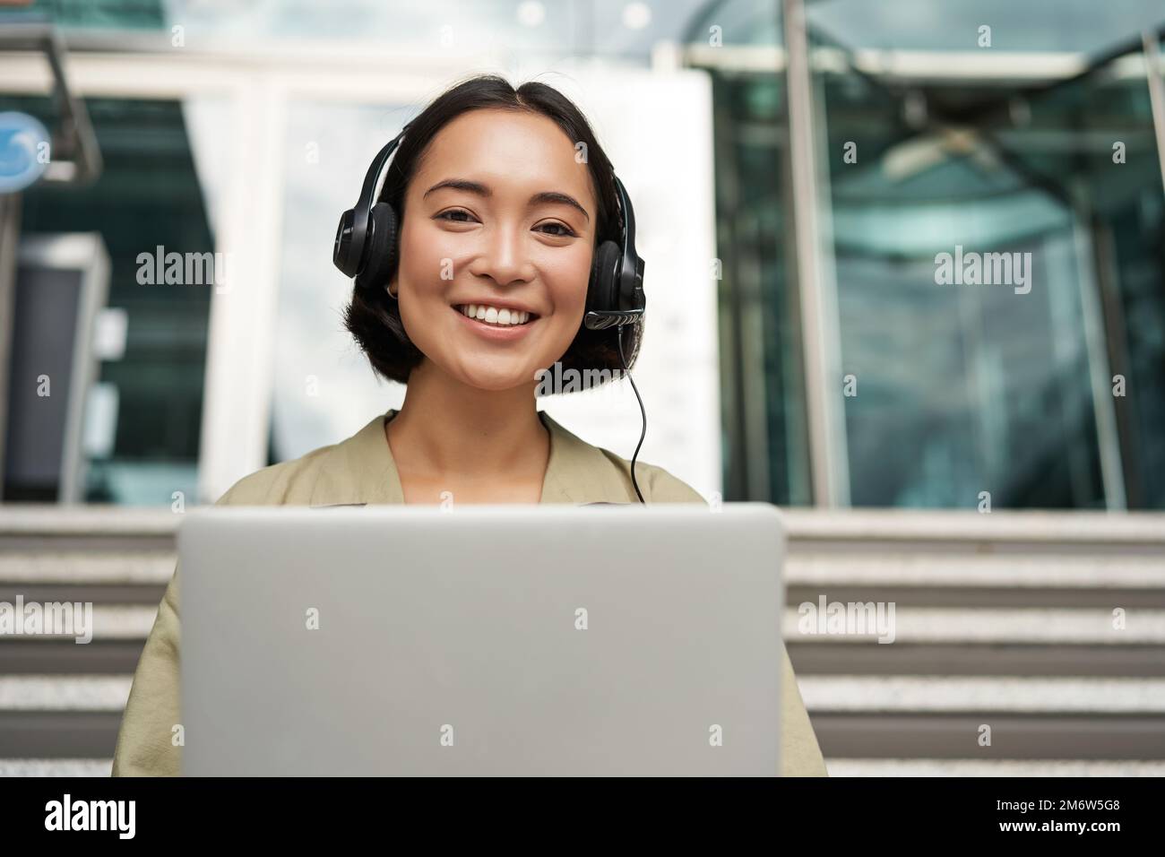 Portrait of young asian woman sitting with laptop and headphones, watching video, does online course on computer, sitting on sta Stock Photo