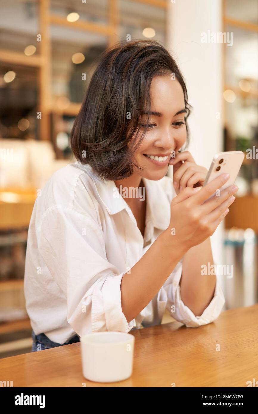 Vertical shot of brunette asian girl sitting in cafe, using smartphone app, chatting on mobile phone and smiling Stock Photo