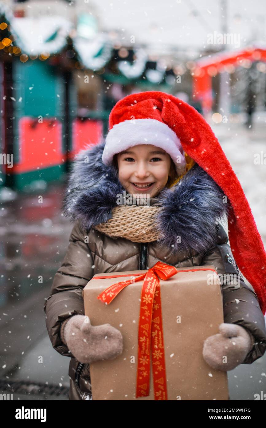 Portrait of joyful girl in Santa hat with gift box for Christmas on city street in winter with snow Stock Photo