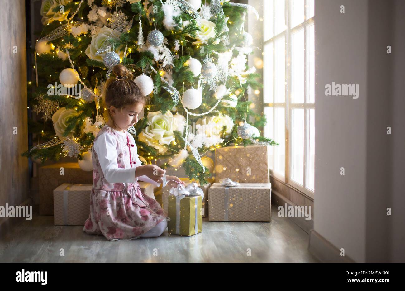 Little girl in beautiful dress is sitting under Christmas tree with gift box  and bow. Light from a l Stock Photo - Alamy