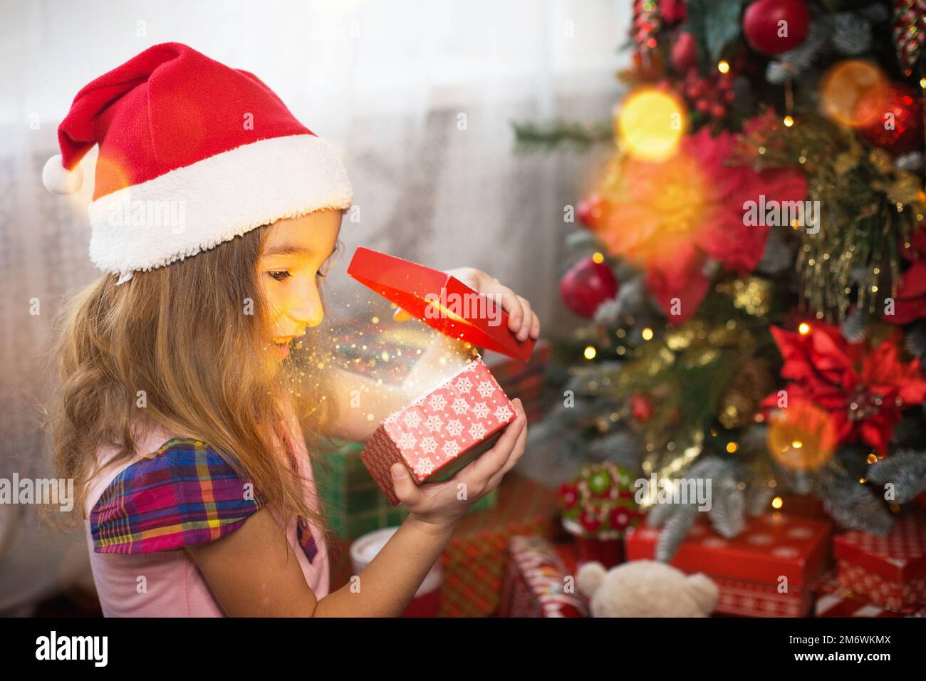 Little girl in Santa hat opens a red box with a gift and a Golden magic light near the Christmas tre Stock Photo