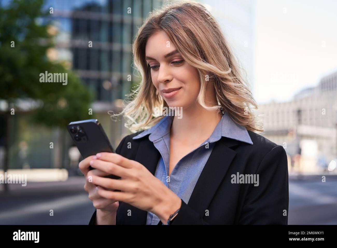 Close up portrait of young corporate woman in black suit, holds smartphone, texting message while standing on street, using mobi Stock Photo