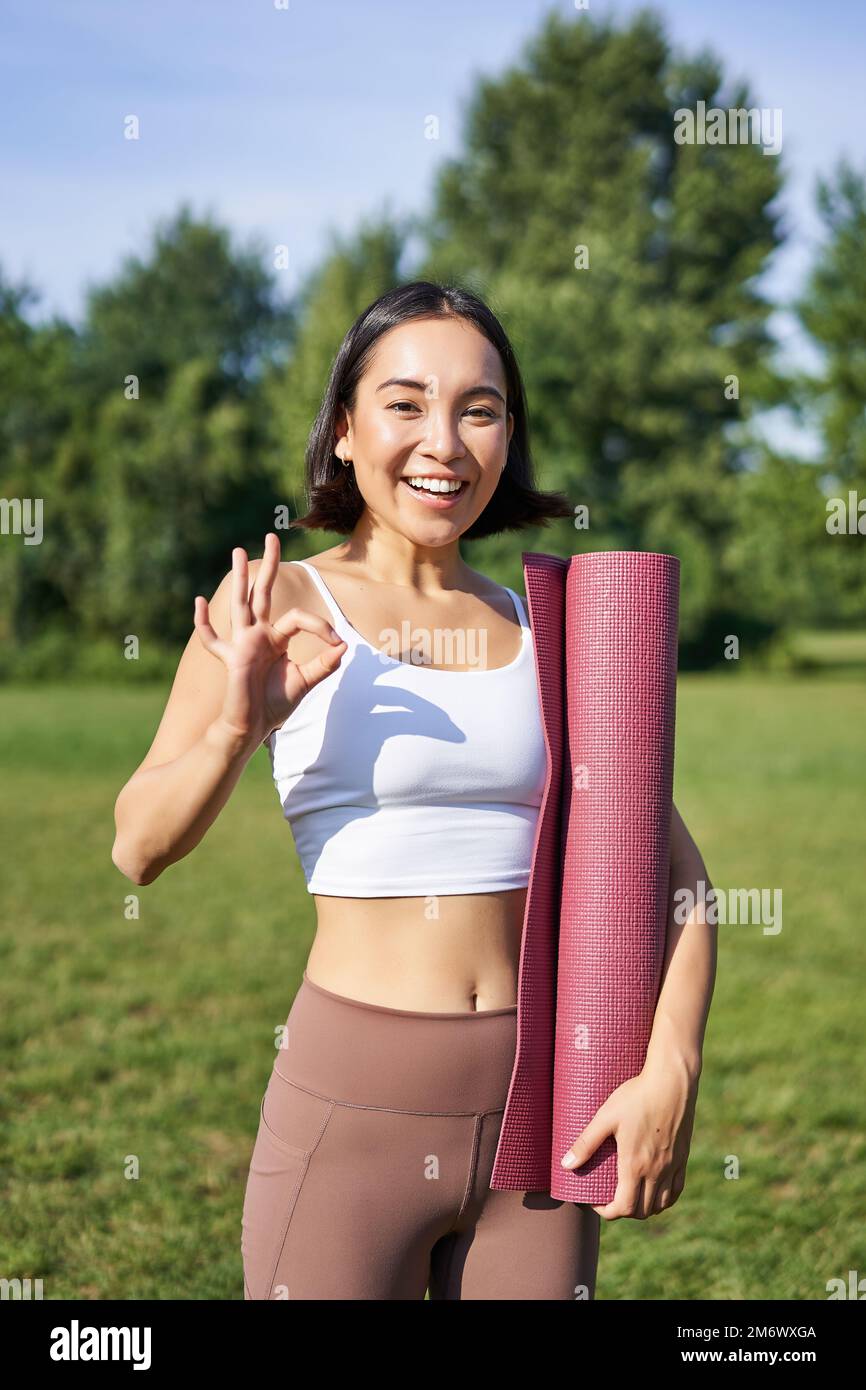 Excited young woman standing with sports mat, yoga clothes, shows okay