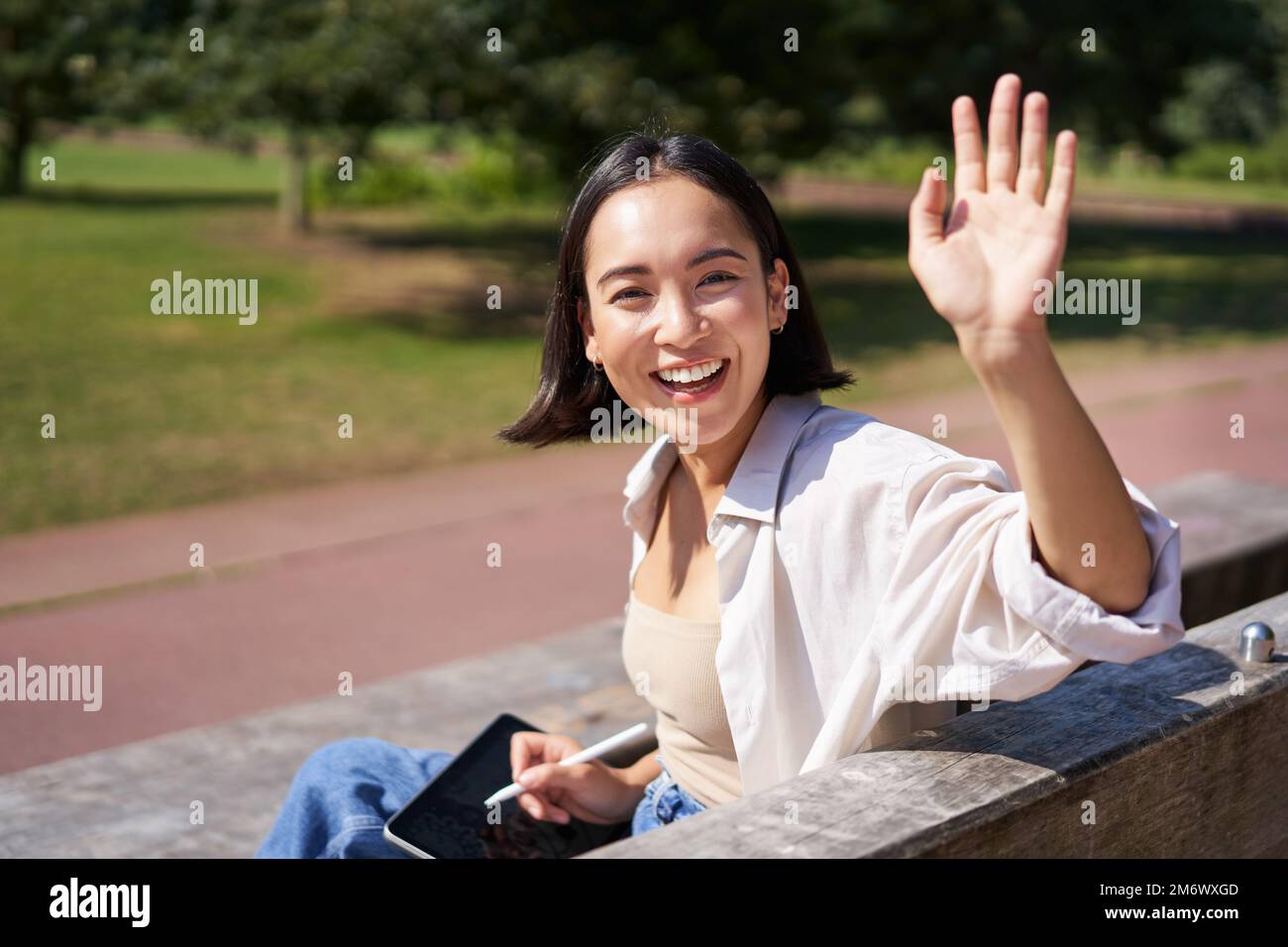 Portrait of happy asian girl say hi, stops drawing on digital tablet with graphic pen and waving hand at you Stock Photo