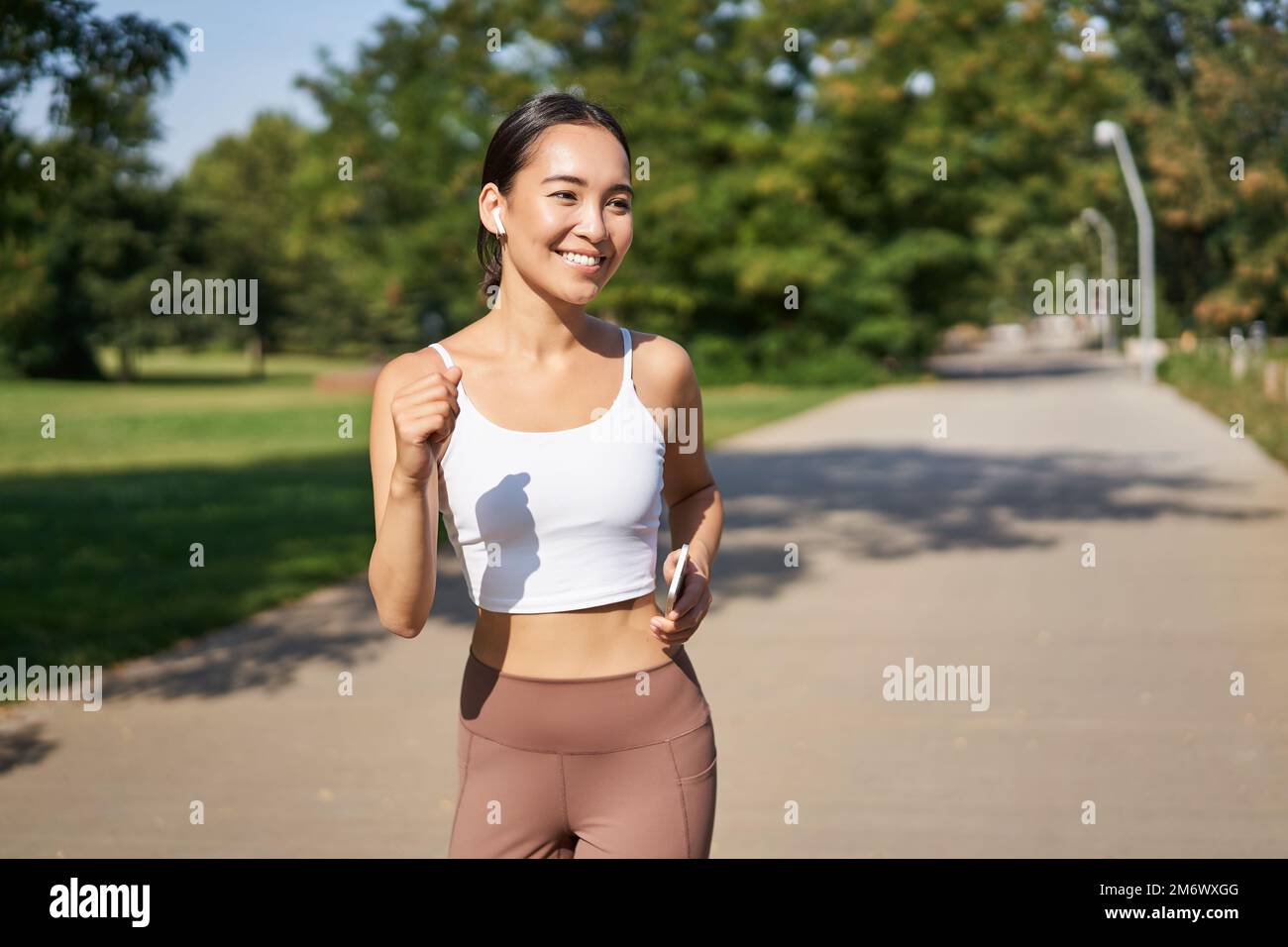Happy smiling asian woman jogging in park. Healthy young female runner doing workout outdoors, running on streets Stock Photo